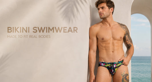 Tattooed model in colorful printed swim briefs beside a seaside arch, next to the words Bikini Swimwear Made to Fit Real Bodies.